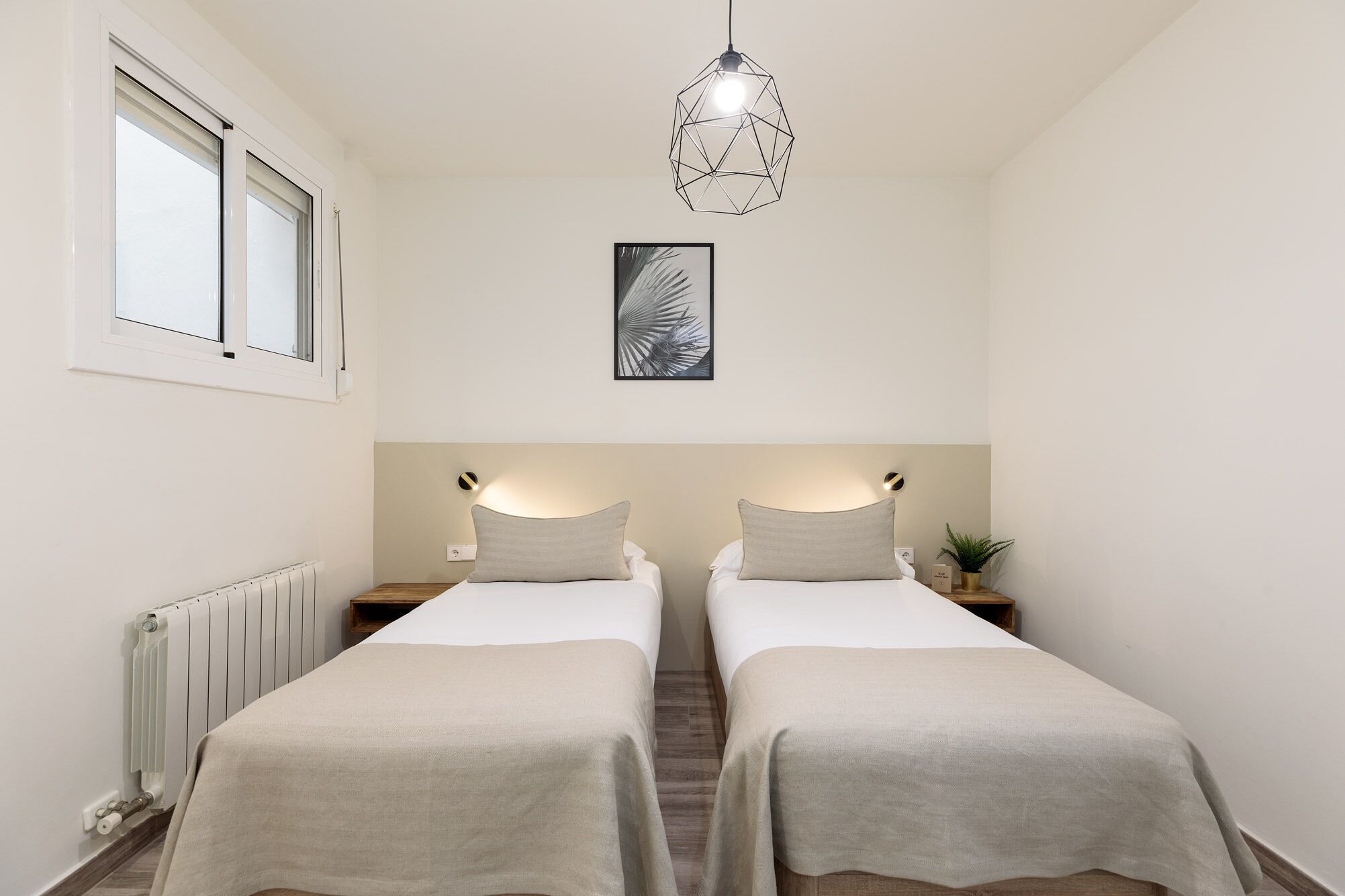 A bedroom with two beds, a radiator, a window, a wall-mounted light, and a geometric pendant light.