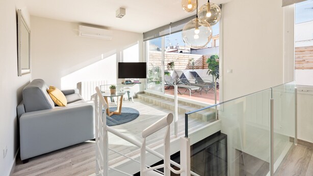 Un salon moderne avec un canapé gris, une table à manger avec des chaises, et une balustrade en verre donnant sur un balcon agrémenté de plantes et d’un téléviseur.