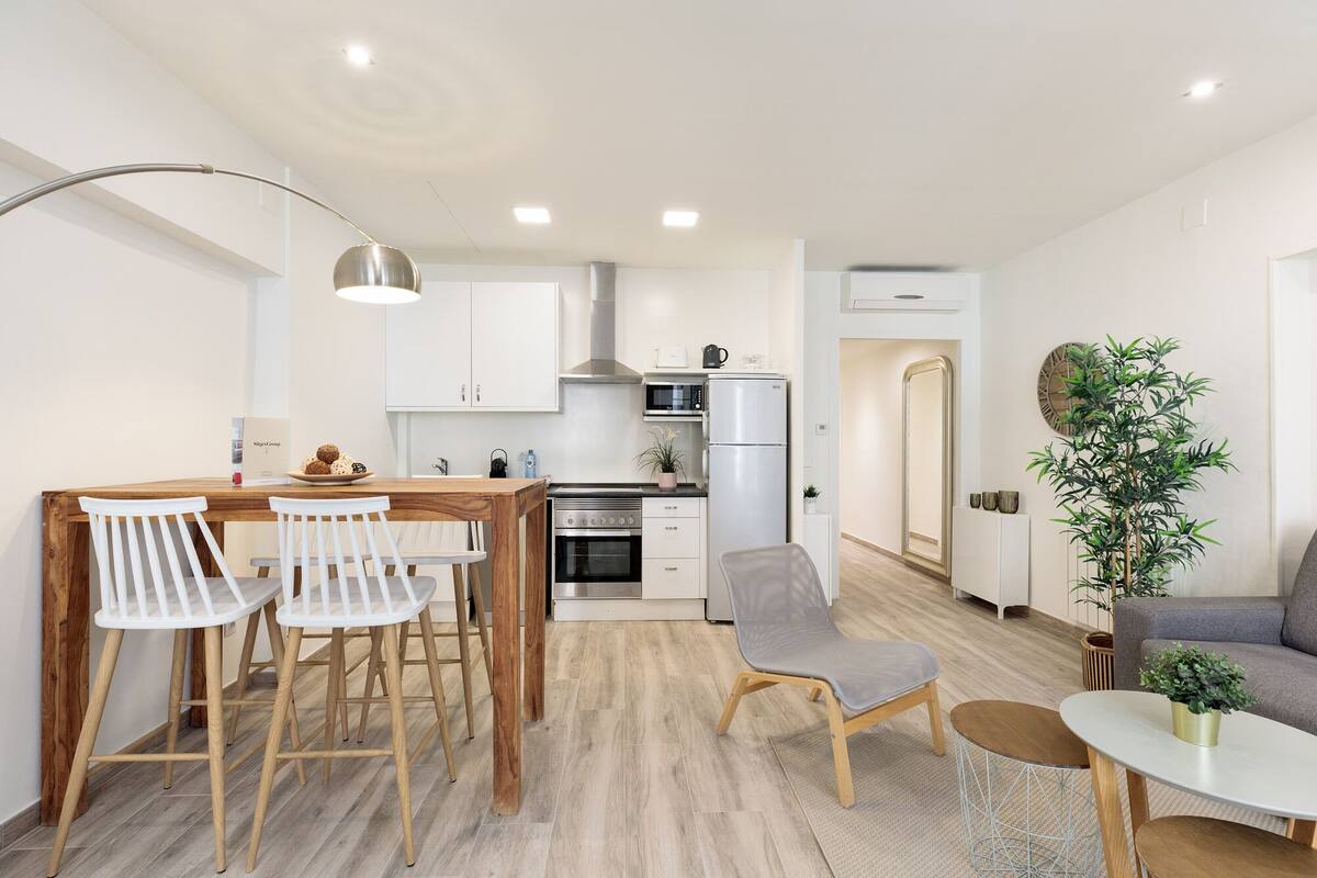A modern kitchen with white cabinets, a stainless steel refrigerator, and a microwave. There is a dining area with a wooden table and white chairs.