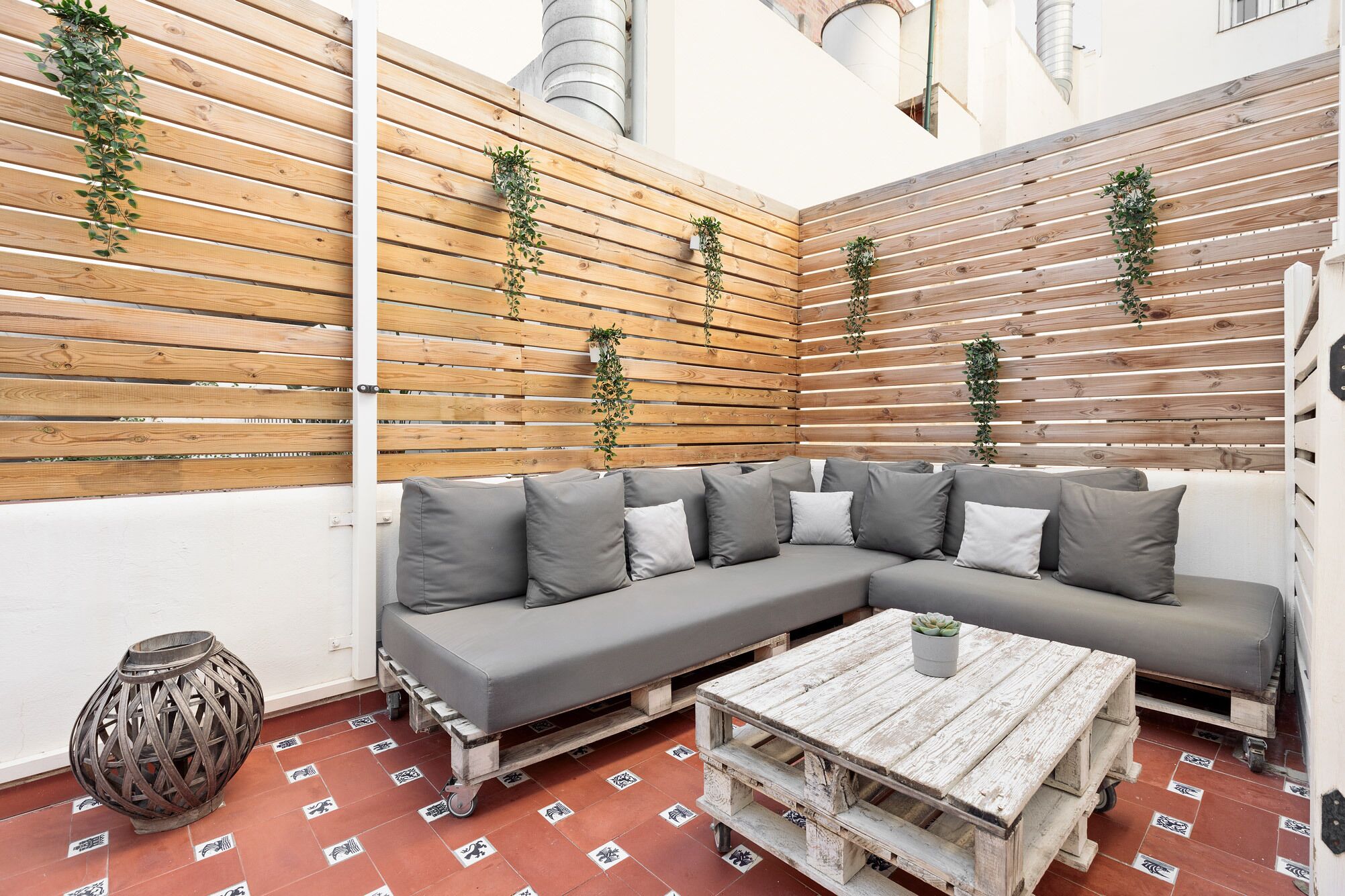 A modern outdoor seating area with a grey sectional sofa, a rustic wooden coffee table, and vertical wooden slats with greenery.
