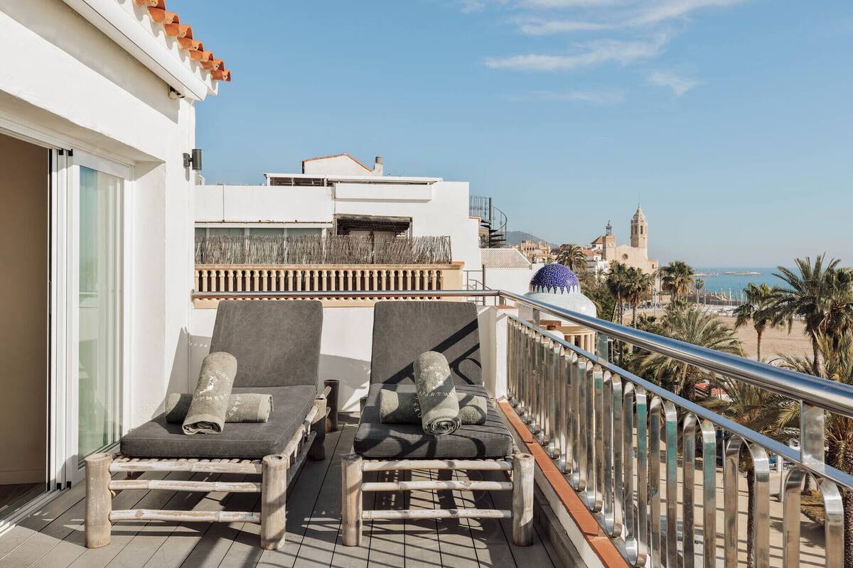 Een balkon met twee loungestoelen, een metalen hekwerk en uitzicht op een strand en gebouwen in de verte.