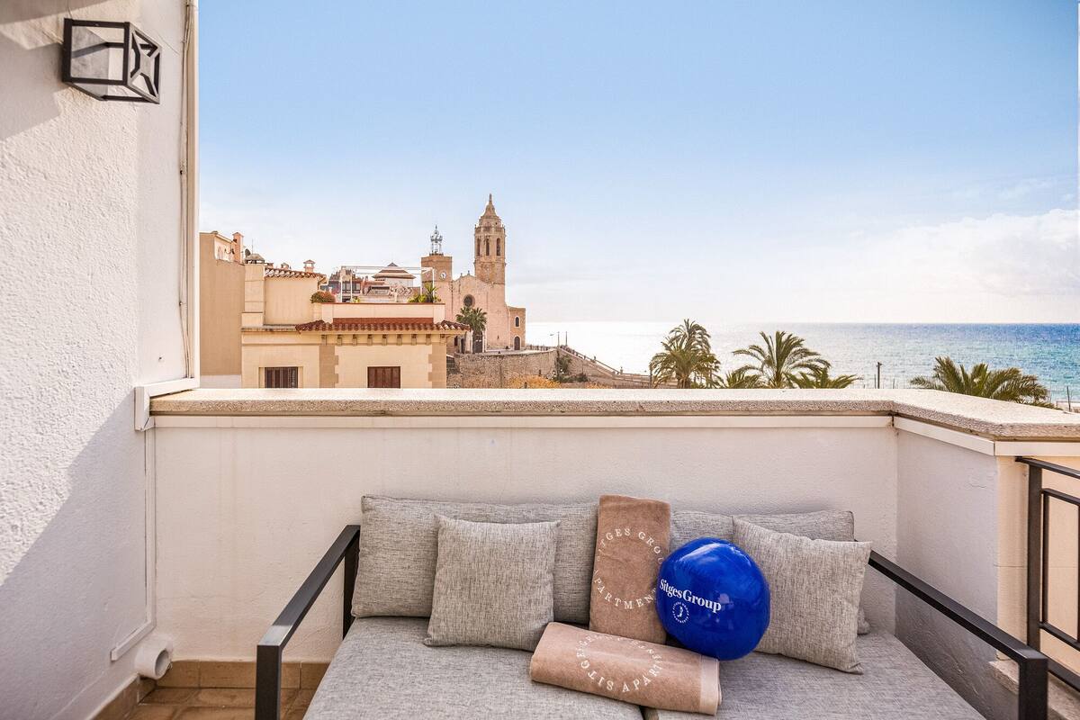 Een balkon met uitzicht op een kustplaatsje, een bank met kussens, en een blauwe ballon.