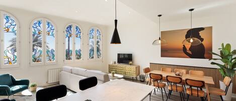 A modern living room with a white sofa, a dining area with wooden chairs, and large windows with stained glass.