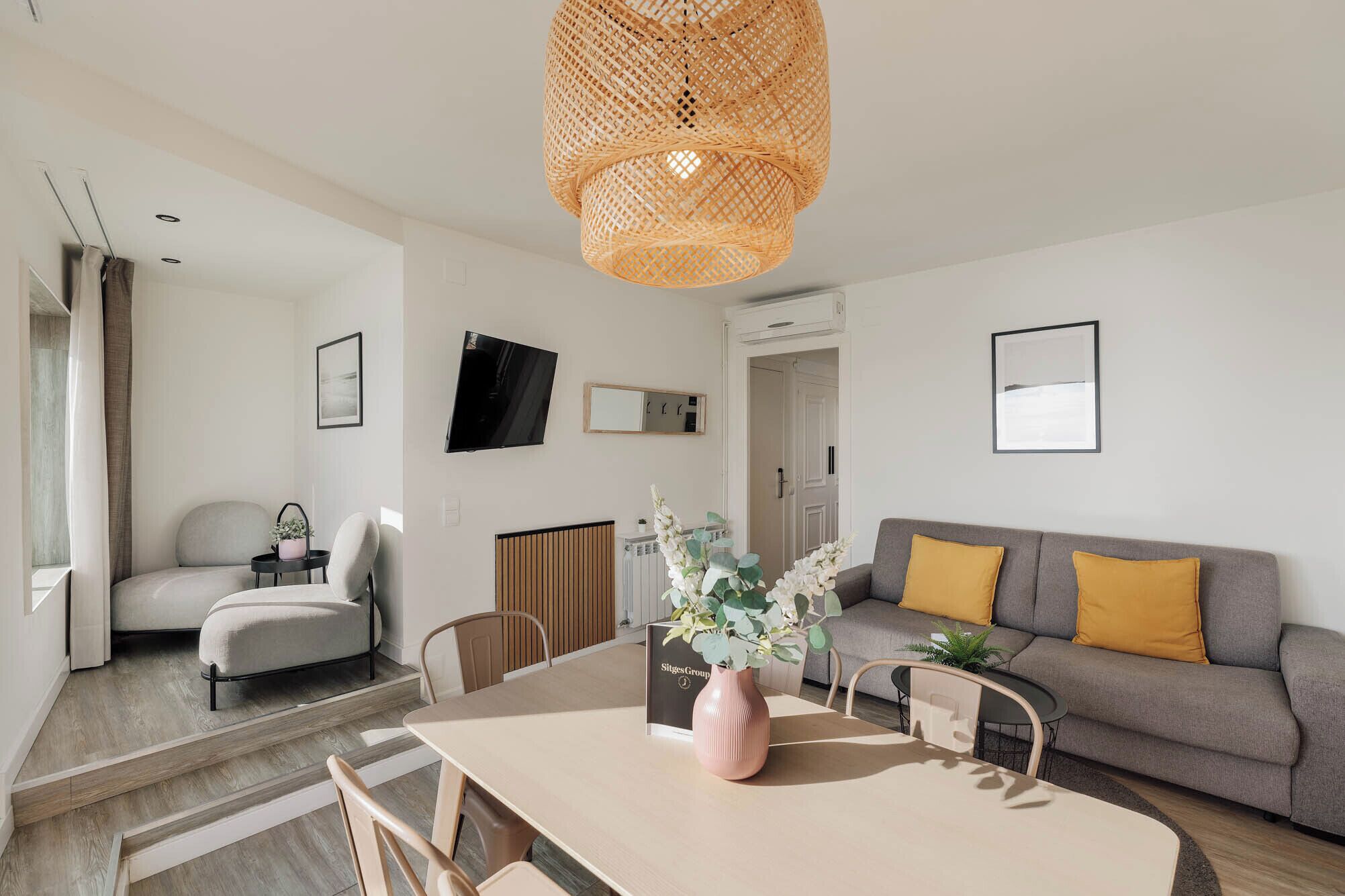 A modern living room with a sofa, a dining table with chairs, a TV mounted on the wall, and a wicker ceiling light.