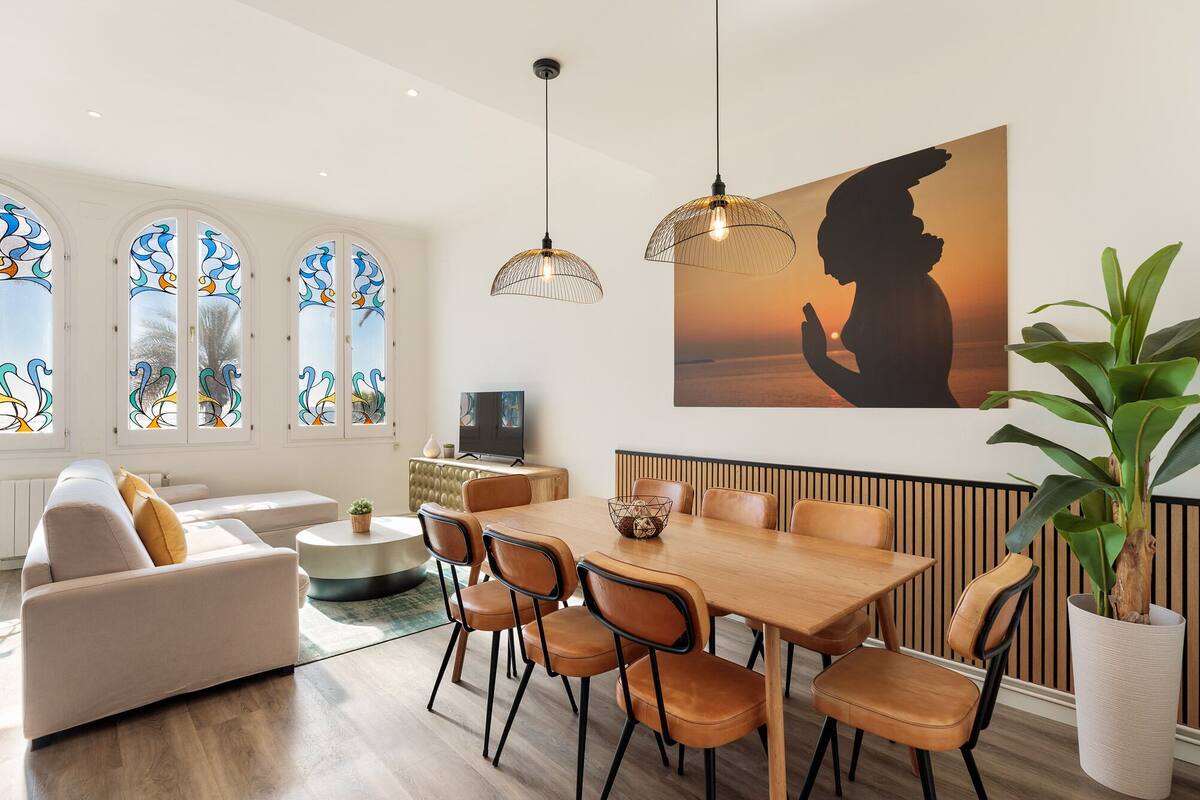 Een moderne woonkamer met een eethoek, met een grote tafel en stoelen, een bank, een televisie en een schilderij van een vrouw aan de muur.