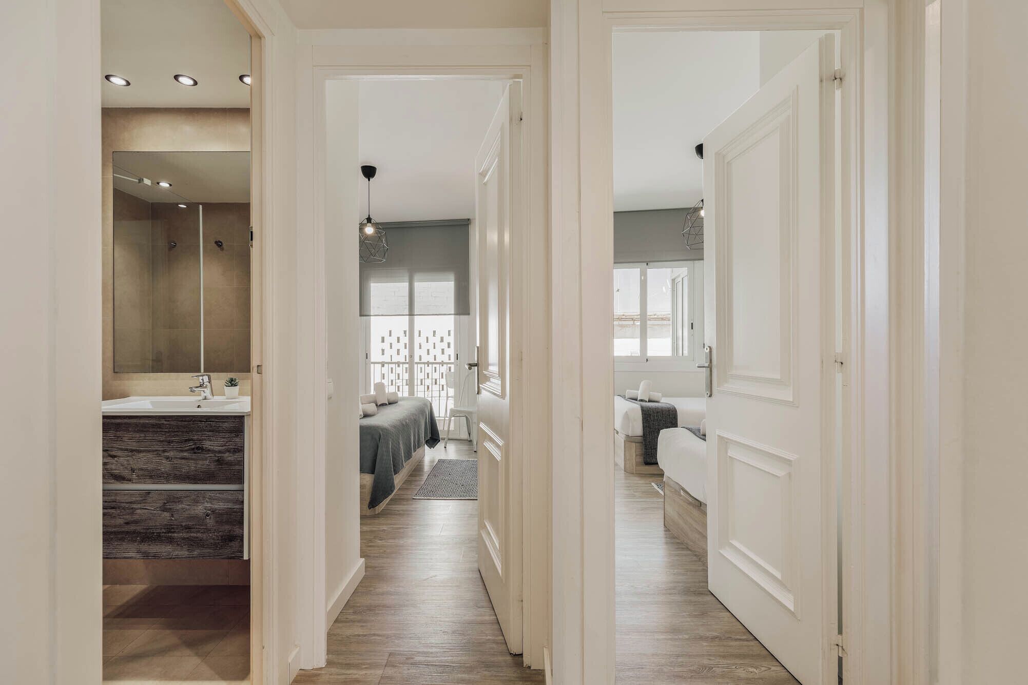 A hallway leading to a bedroom with a large window, a bathroom with a mirror and sink, and a wooden floor.