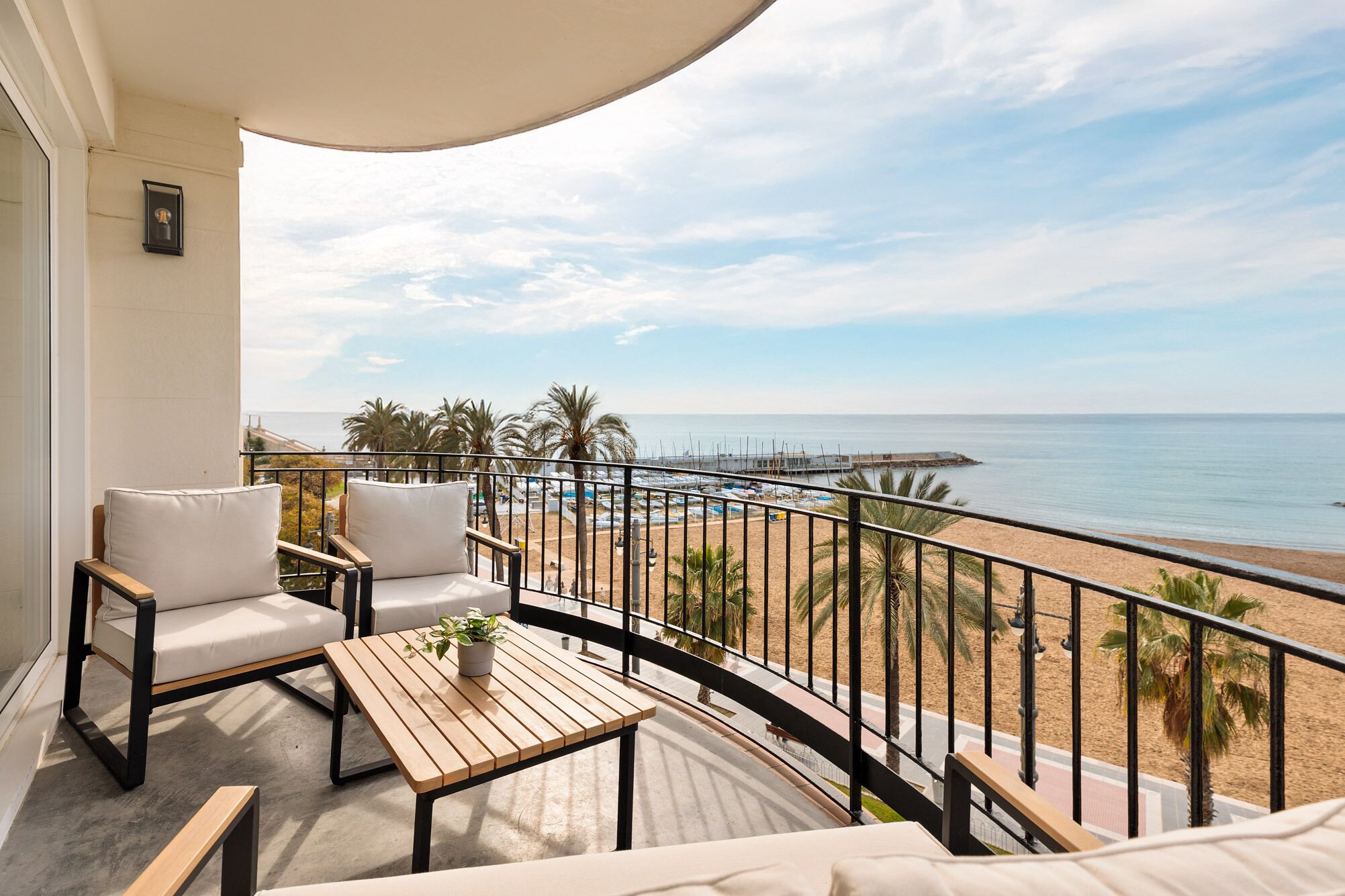 Un balcón con vista a la playa, una mesa de madera y dos sillas blancas con cojines.