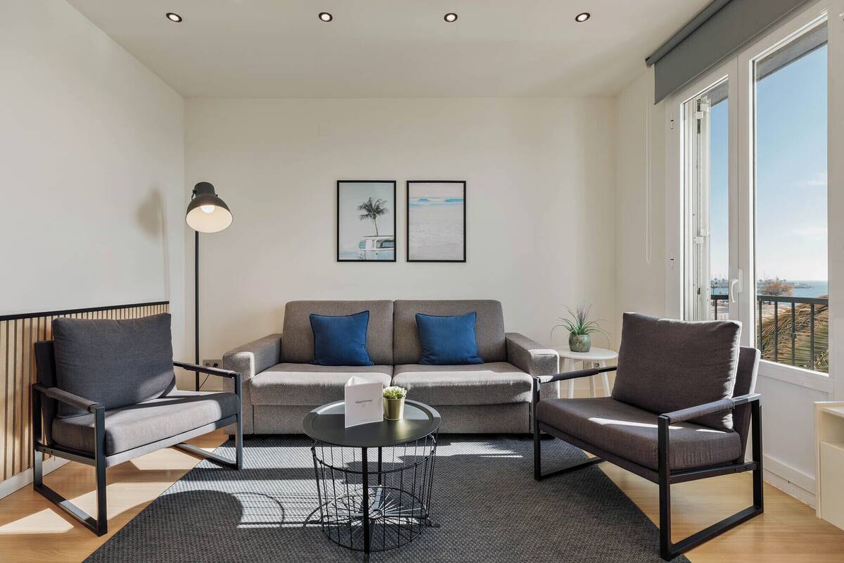 A modern living room with a grey sofa, two armchairs, a round coffee table, and a floor lamp. There are two framed pictures on the wall and a window with a view of the city.