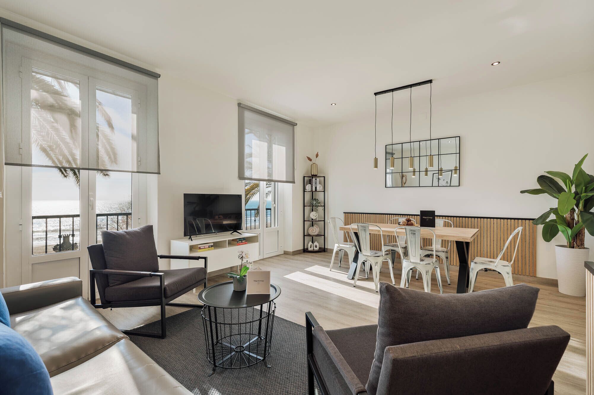 Ein modernes Wohnzimmer mit Sofa, Sesseln, Fernseher, Essbereich mit Tisch und Stühlen, Bücherregal und großem Fenster mit Aussicht.