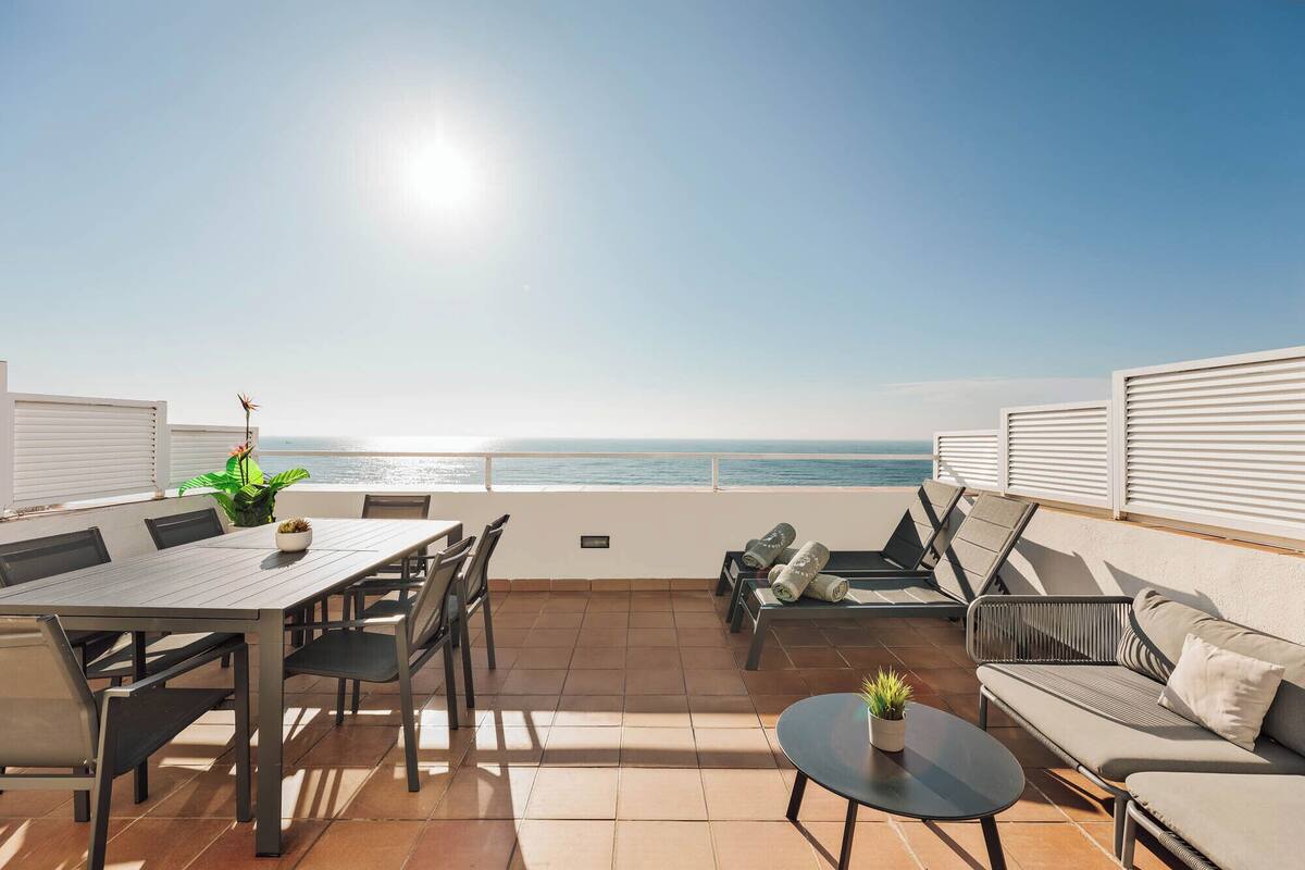 A terrace with a table and chairs, a sofa, and a small table with a plant, overlooking the sea.