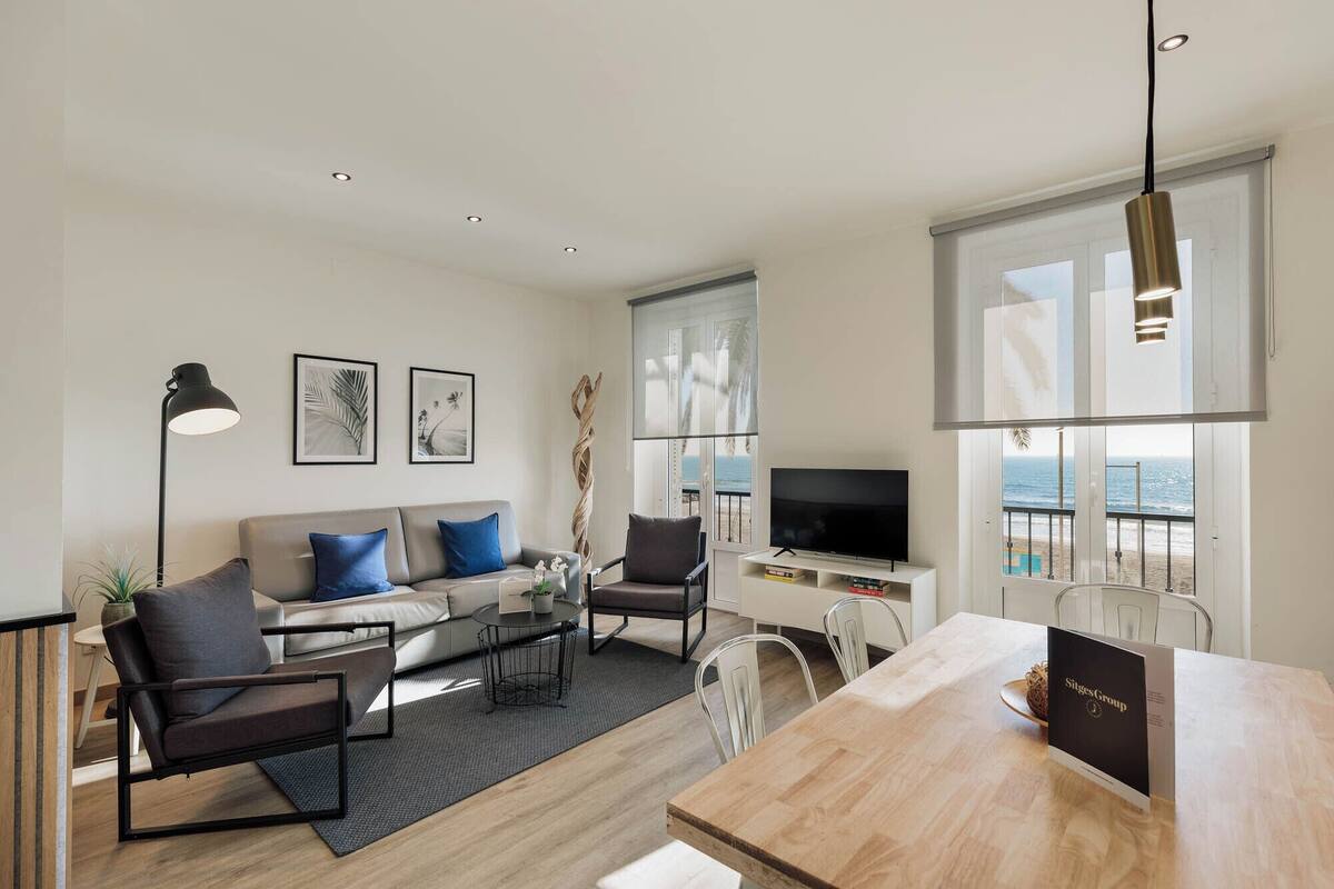 A modern living room with a sofa, coffee table, and chairs, a dining table with a sign, a TV, and a large window with a view of the beach.