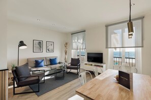 A modern living room with a sofa, coffee table, and chairs, a dining table with a sign, a TV, and a large window with a view of the beach. - Sitges Group Beach Dreams (Sitges)