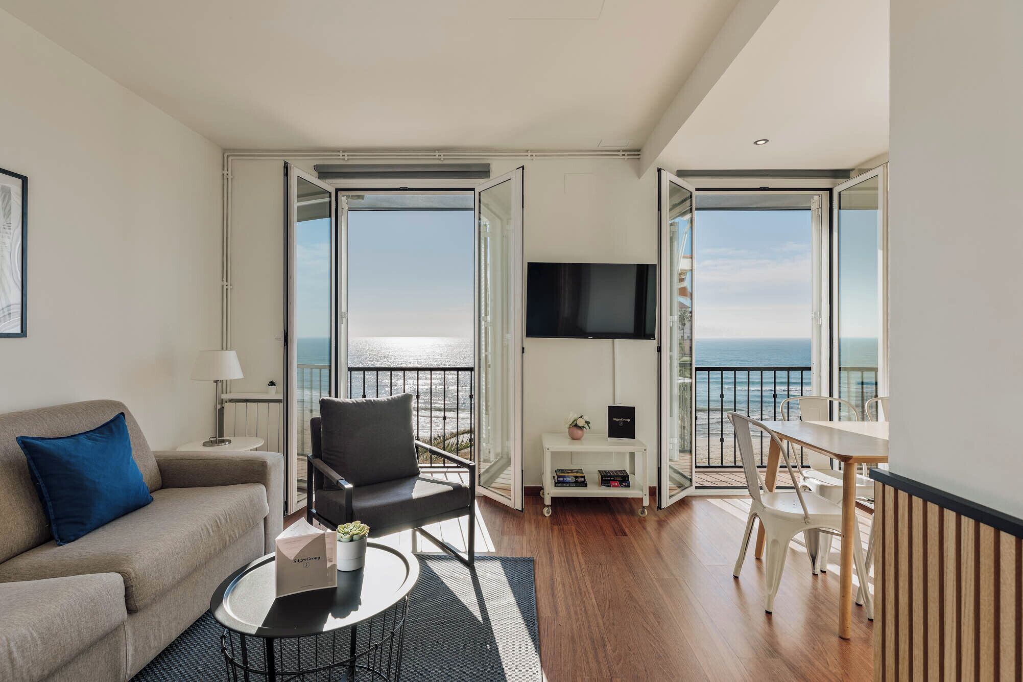 A modern living room with a sofa, a coffee table, and a TV. There is a balcony with a view of the sea.
