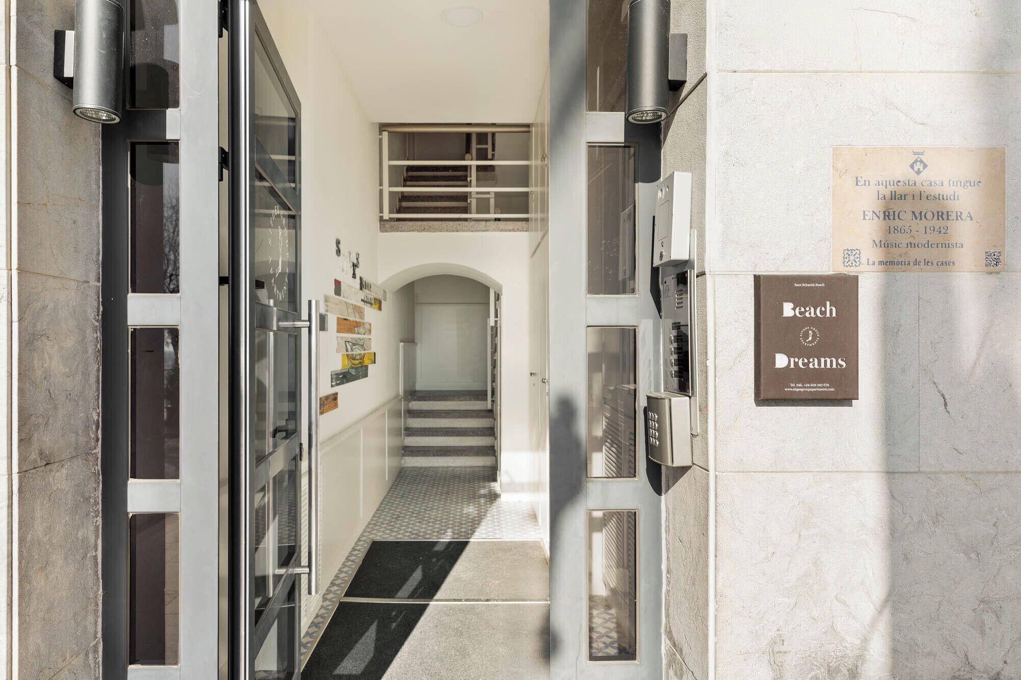 A hallway with a staircase leading to a landing, a plaque commemorating Enrico Morera, and a sign for Beach Dreams.