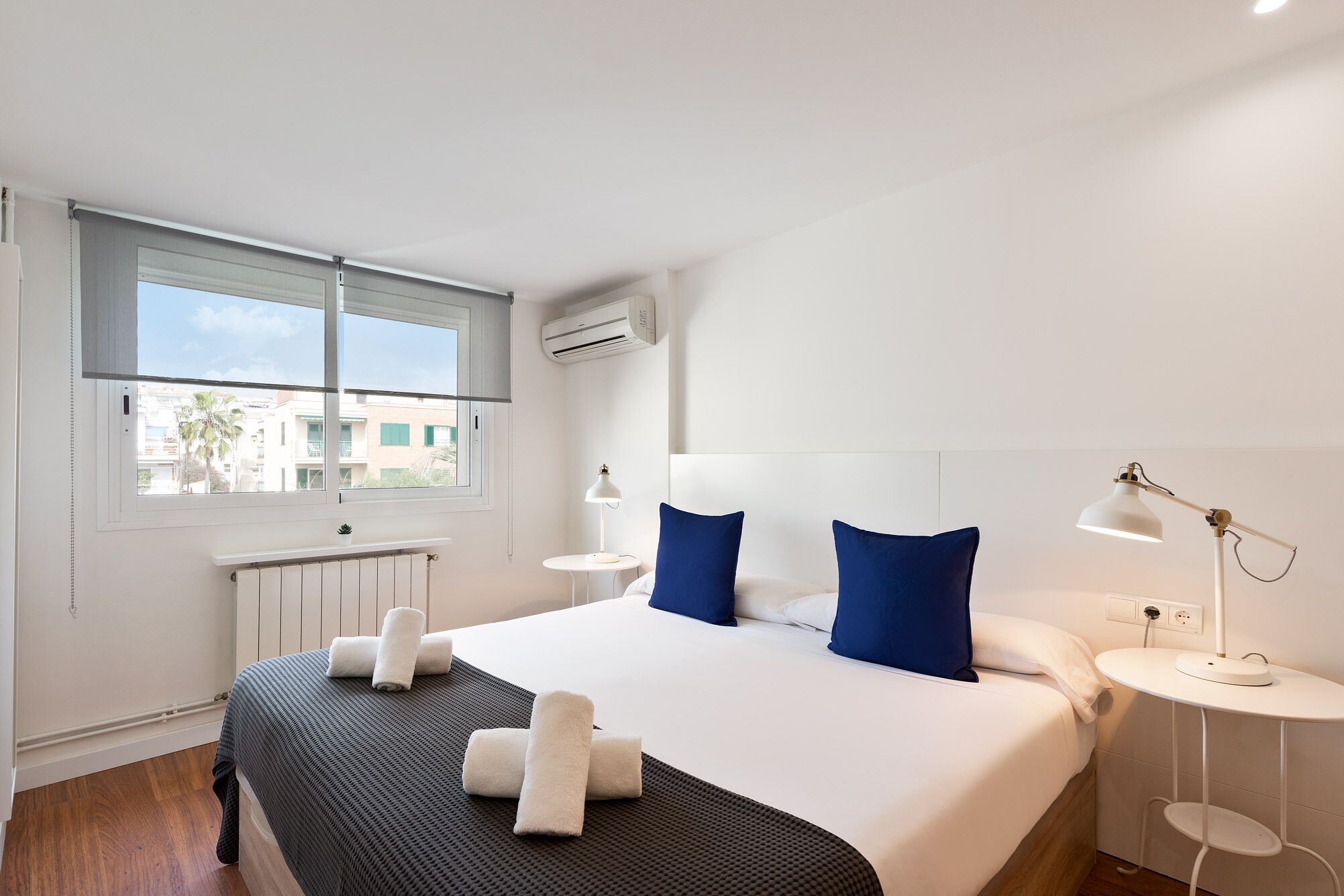 A hotel room with a large bed, two bedside tables, a window with a view of buildings, and an air conditioning unit.