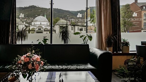 A room with a view of a building with domes, a black leather sofa, a table with a vase of flowers, and several potted plants. - Hotel President Sarajevo (Sarajevo)