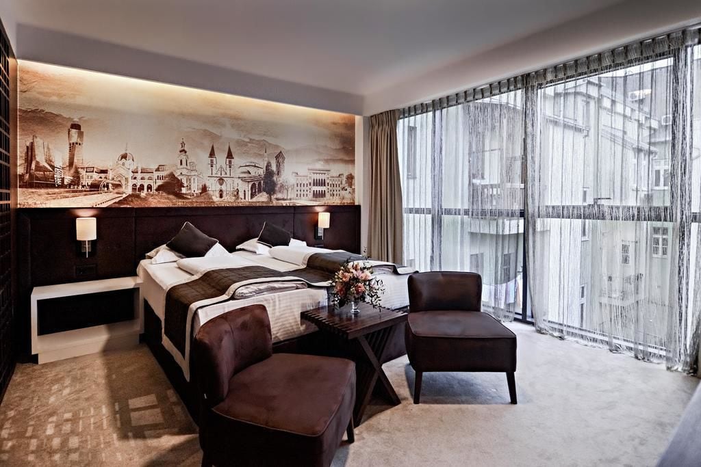 A hotel room with a large bed, a desk with a lamp, two chairs, and a large window with curtains.