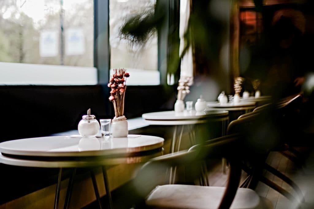 A restaurant with round tables and chairs, a vase with dried flowers, and a window with a view of trees.