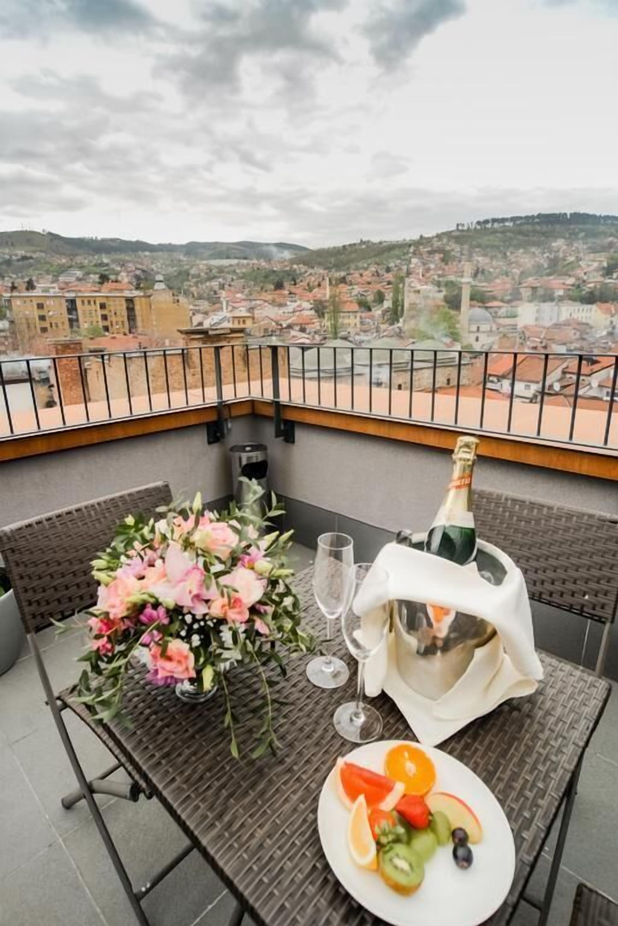 A balcony with a table set for two, featuring a bouquet of flowers, a bottle of champagne, and a plate of fruit.