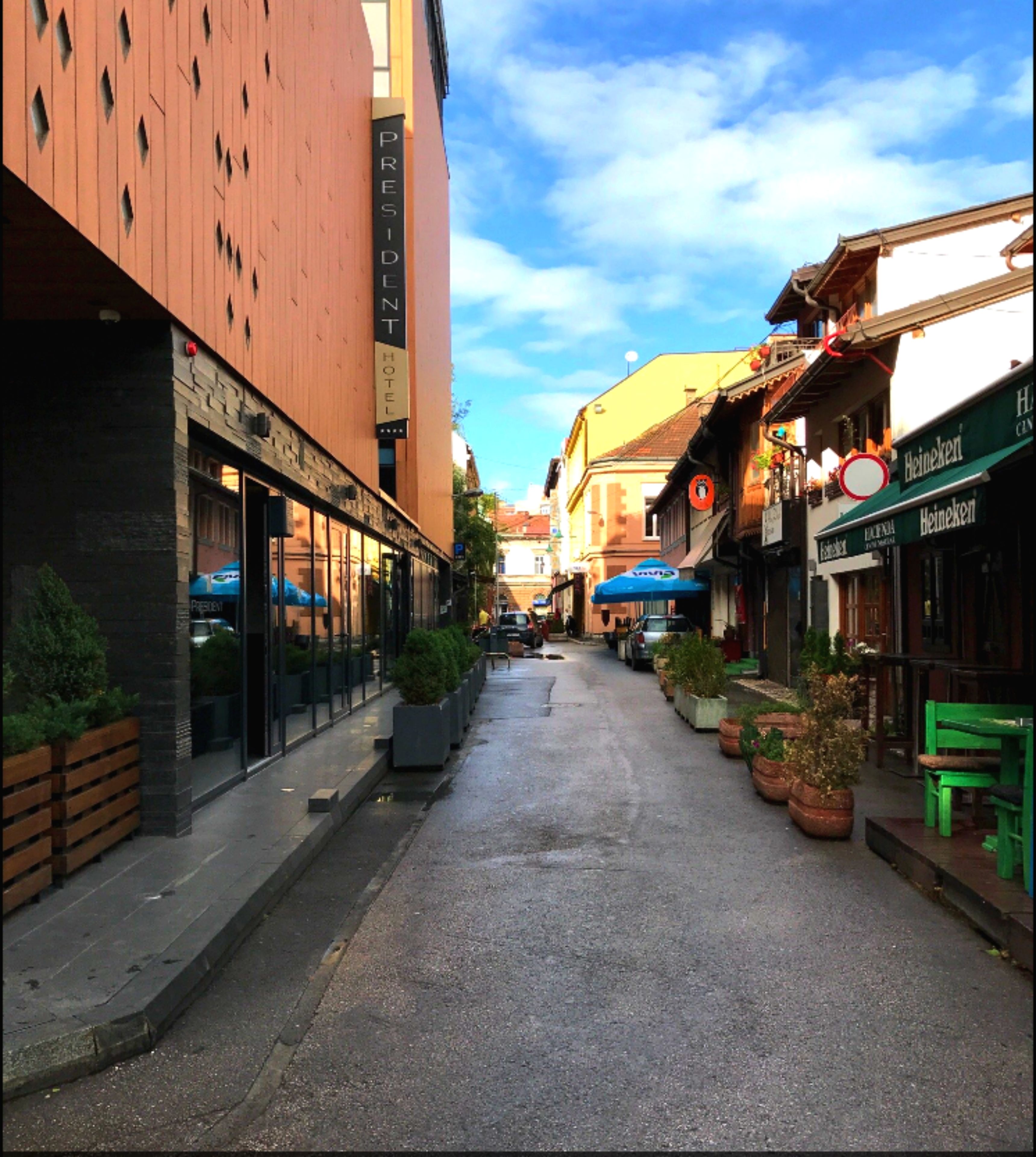 En smal gata med byggnader på båda sidor, inklusive ett hotell som heter ”President Hotel” och en Heineken-bar. Gatan är kantad med planterade blommor och det finns en bänk på höger sida.