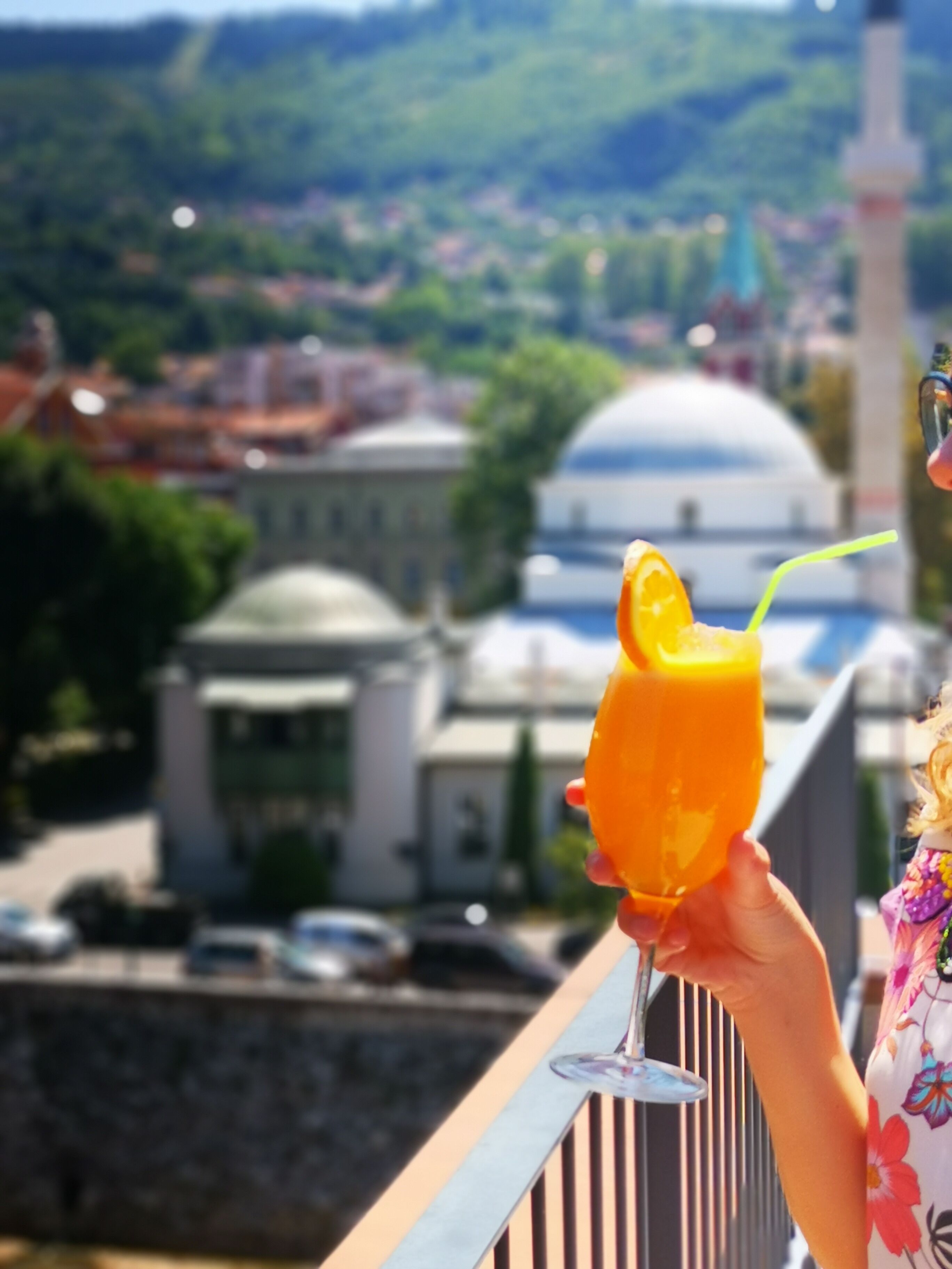A person holding a glass of orange juice with a straw, overlooking a cityscape with buildings and a dome structure.