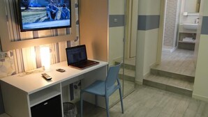 Une chambre d’hôtel équipée d’un bureau, d’un ordinateur portable, d’une télévision et d’une salle de bain visible à travers une paroi en verre.