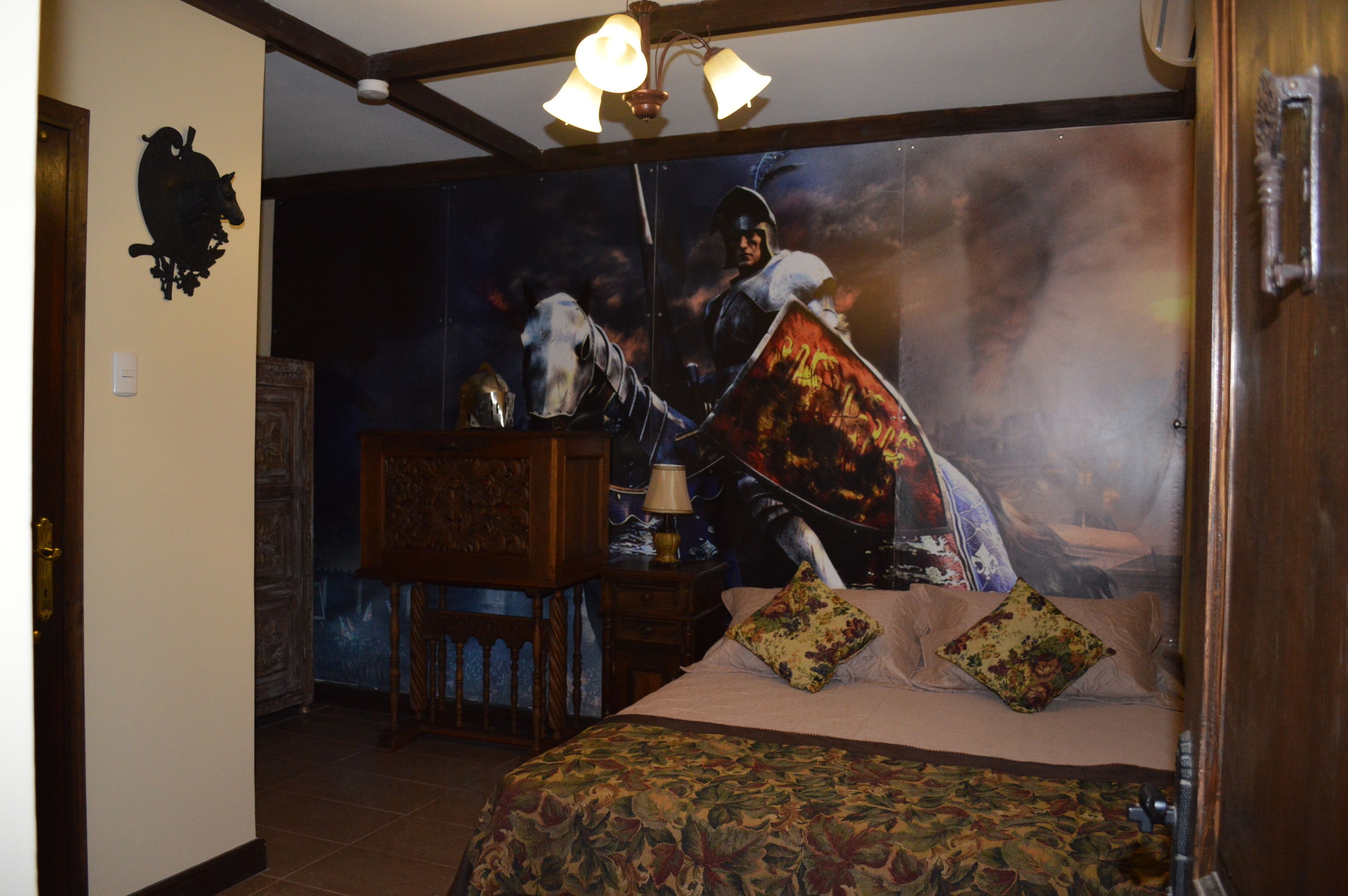 A bedroom with a large bed, a wooden nightstand, a lamp, and a tapestry depicting a knight on horseback.