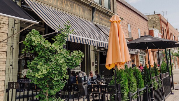 A brick building with a sign reading 'Foster's Inn.' There are outdoor tables and chairs, and a row of potted plants along the sidewalk.