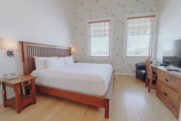 A bedroom with a wooden bed, bedside table with a phone, two windows with blinds, a desk with a chair, and a television.