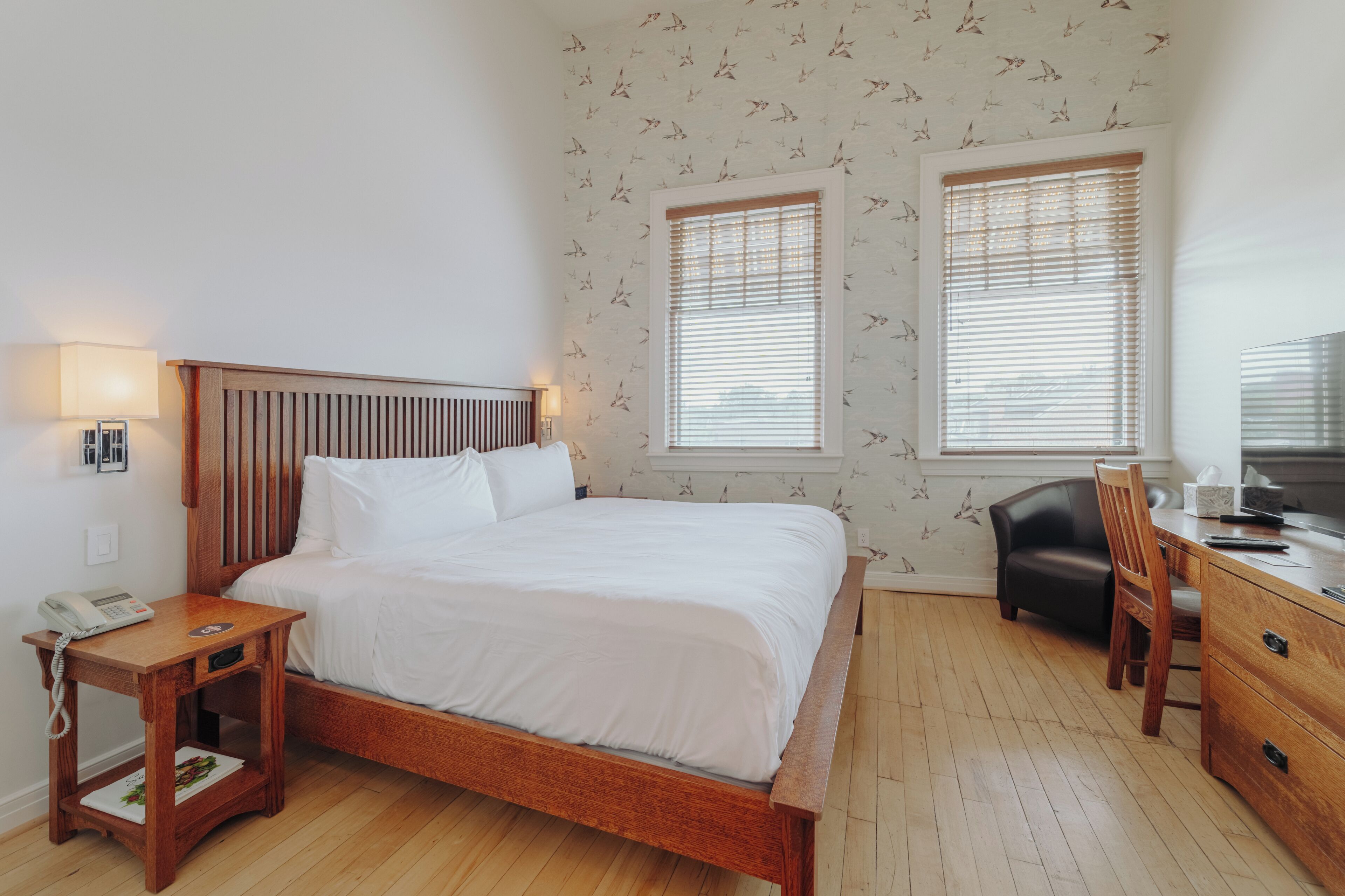 Un dormitorio con cama de madera, mesita de noche con lámpara, dos ventanas con persianas, un escritorio con silla y un papel tapiz estampado.