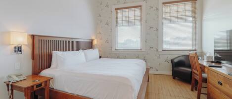 Un dormitorio con cama de madera, mesita de noche con lámpara, dos ventanas con persianas, un escritorio con silla y un papel tapiz estampado.