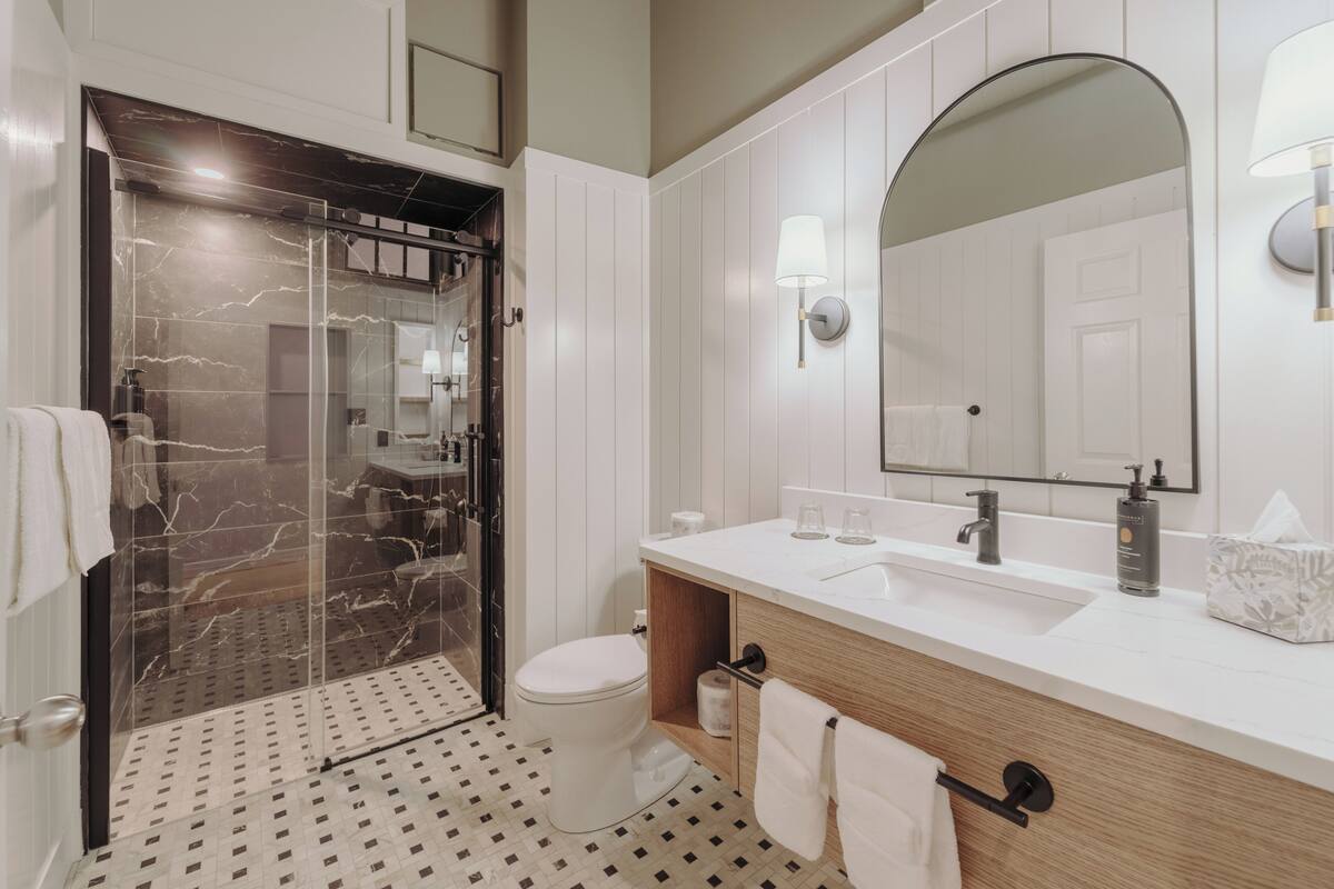 A modern bathroom with a glass-enclosed shower, a white toilet, a wooden vanity with a sink, and a large mirror.