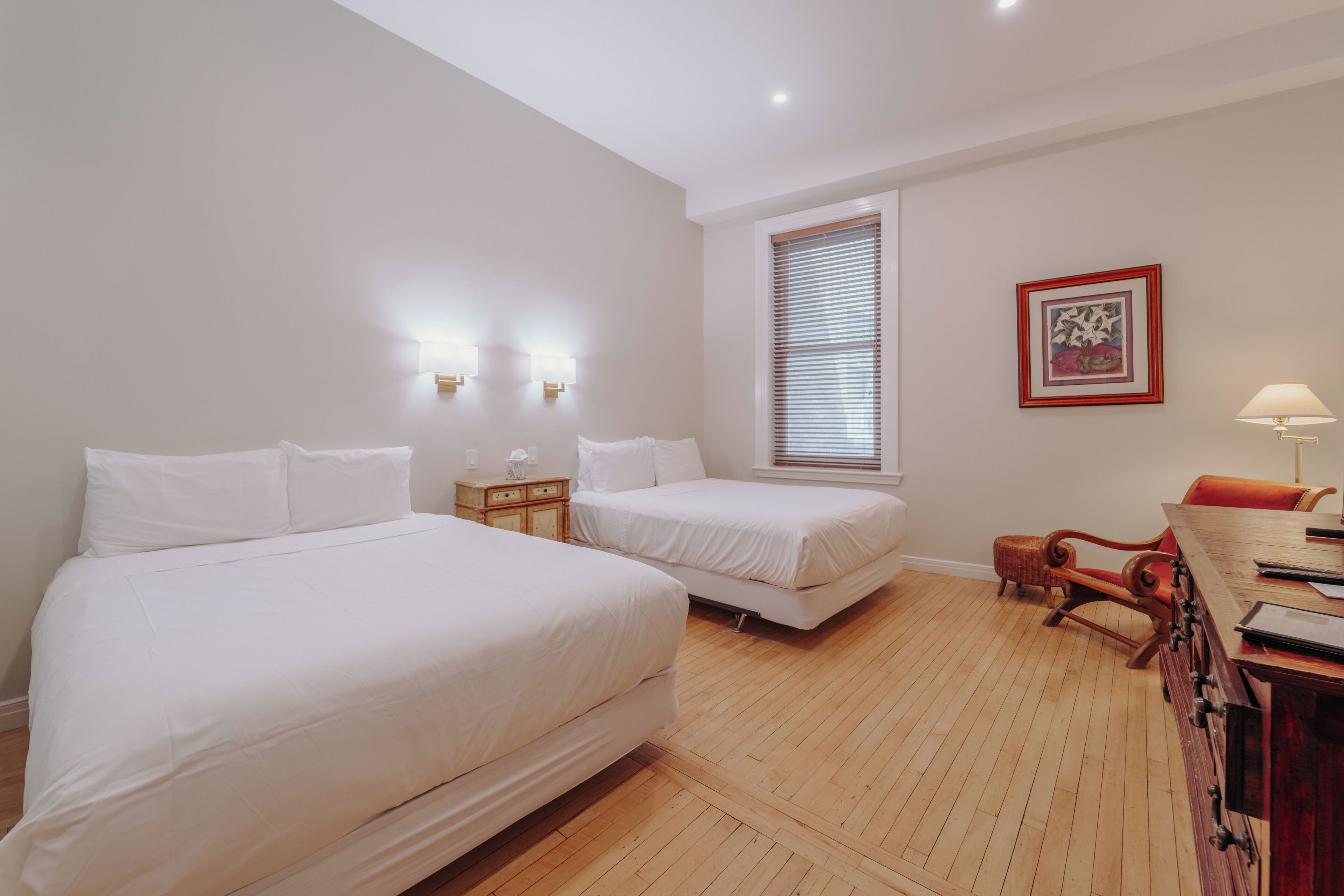 A hotel room with two beds, a wooden floor, a desk with a chair, and a window with blinds.