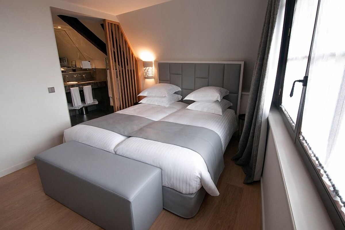 A modern bedroom with a large bed, a bedside table, a built-in bench, and a window with curtains.