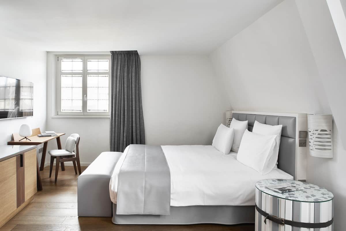 Ein modernes Hotelzimmer mit Bett, Schreibtisch, Stuhl, rundem Beistelltisch und einem Fenster mit Vorhängen.