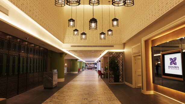 Een hotelgang met behang, hangende kroonluchters, een receptie en een zichtbare winkel genaamd 'Estadia by Hatten'.