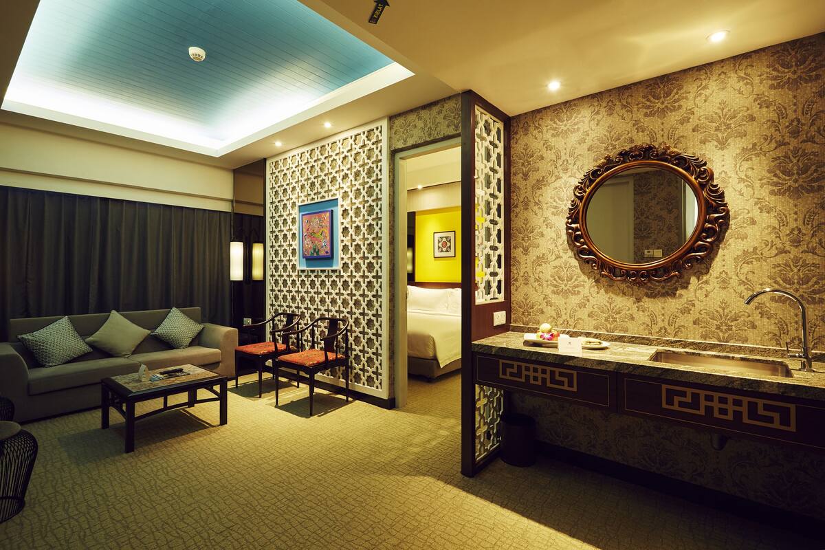 A hotel room with a sofa, chairs, a coffee table, a bed, a bathroom with a sink and mirror, and a decorative wall.