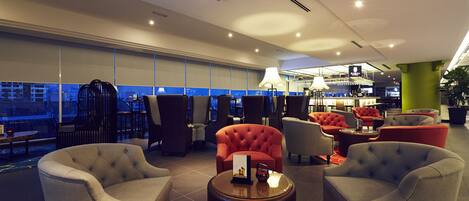 A modern lounge area with comfortable seating, a bar, and large windows offering a city view.