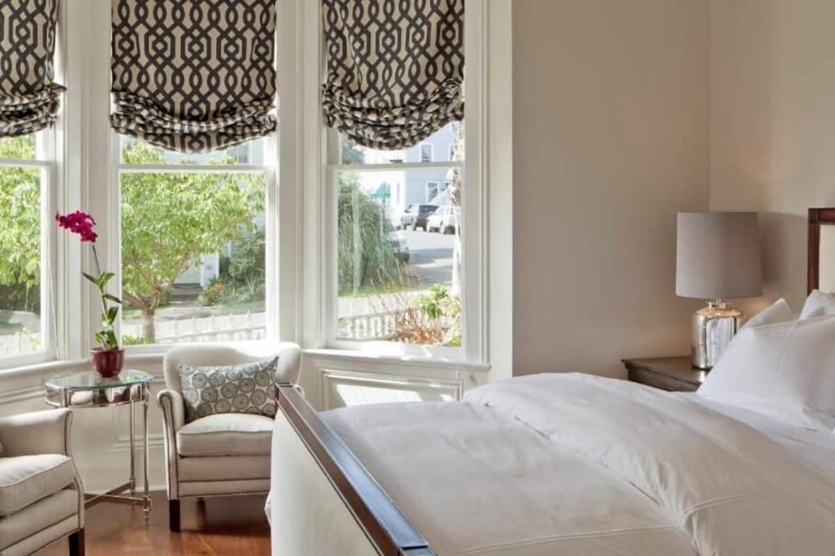 Un dormitorio con una cama grande, una mesita de noche con lámpara, una silla y un ventanal con cortinas estampadas.