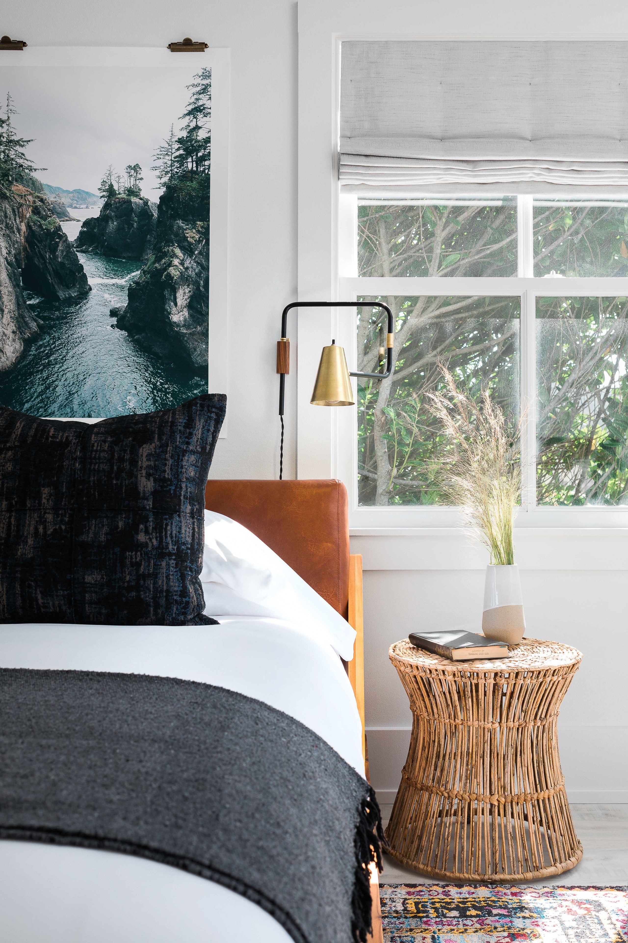 Une chambre à coucher avec un lit, une grande fenêtre donnant sur des arbres, une lampe de chevet et une table d’appoint en osier sur laquelle se trouvent un livre et un vase.