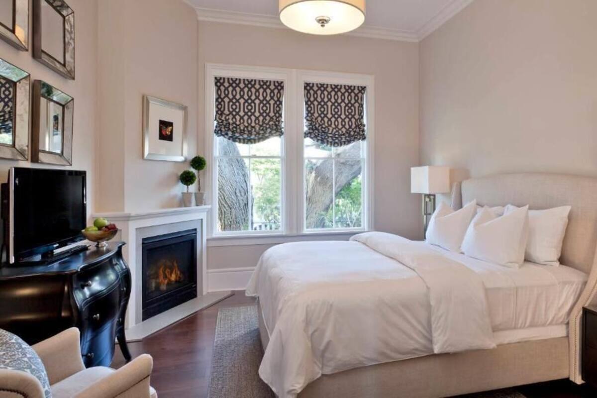Un dormitorio con cama, lámparas de noche, chimenea, televisor y ventana con cortinas.