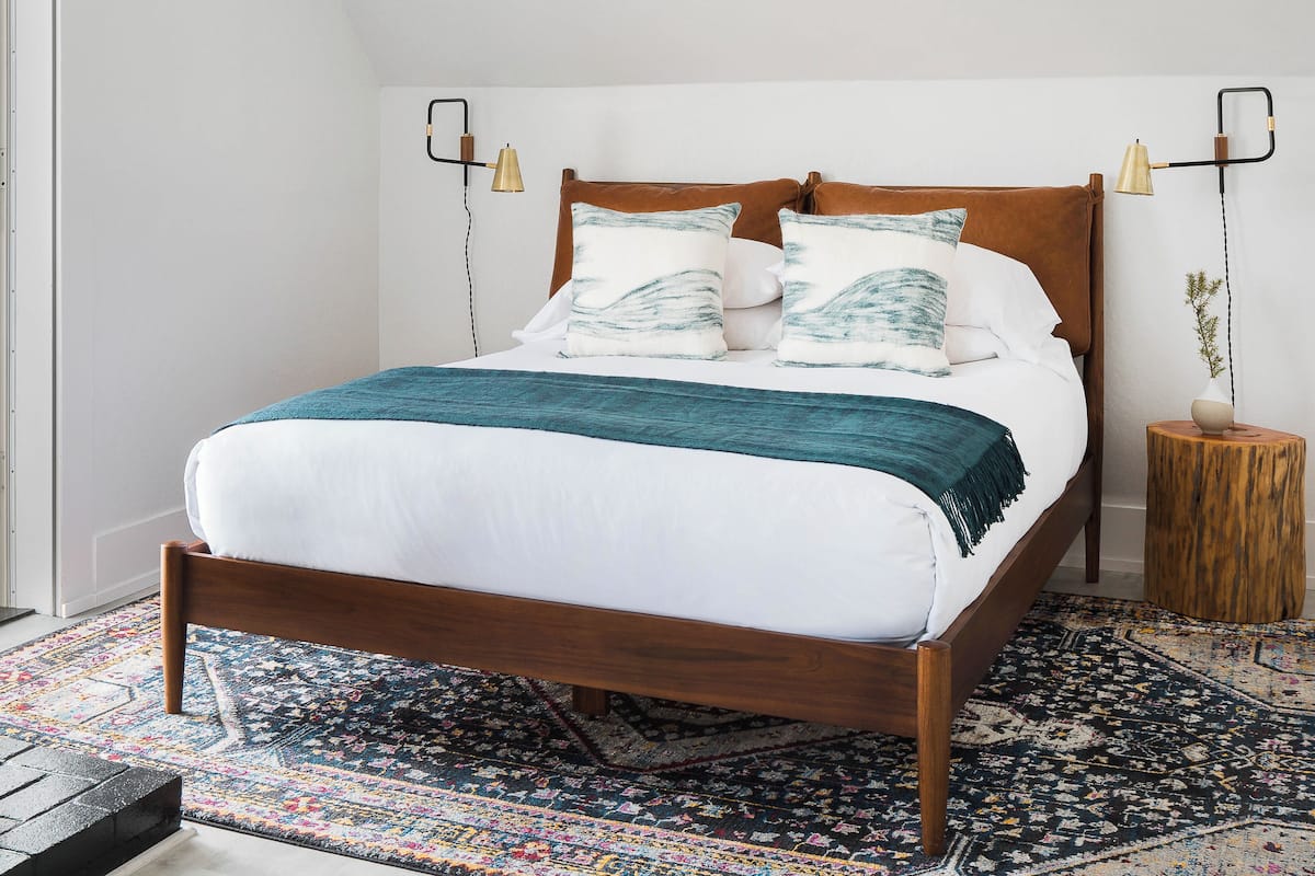 Una cama con estructura de madera, ropa de cama blanca y almohadas estampadas. Junto a la cama, un taburete de madera con un jarrón.
