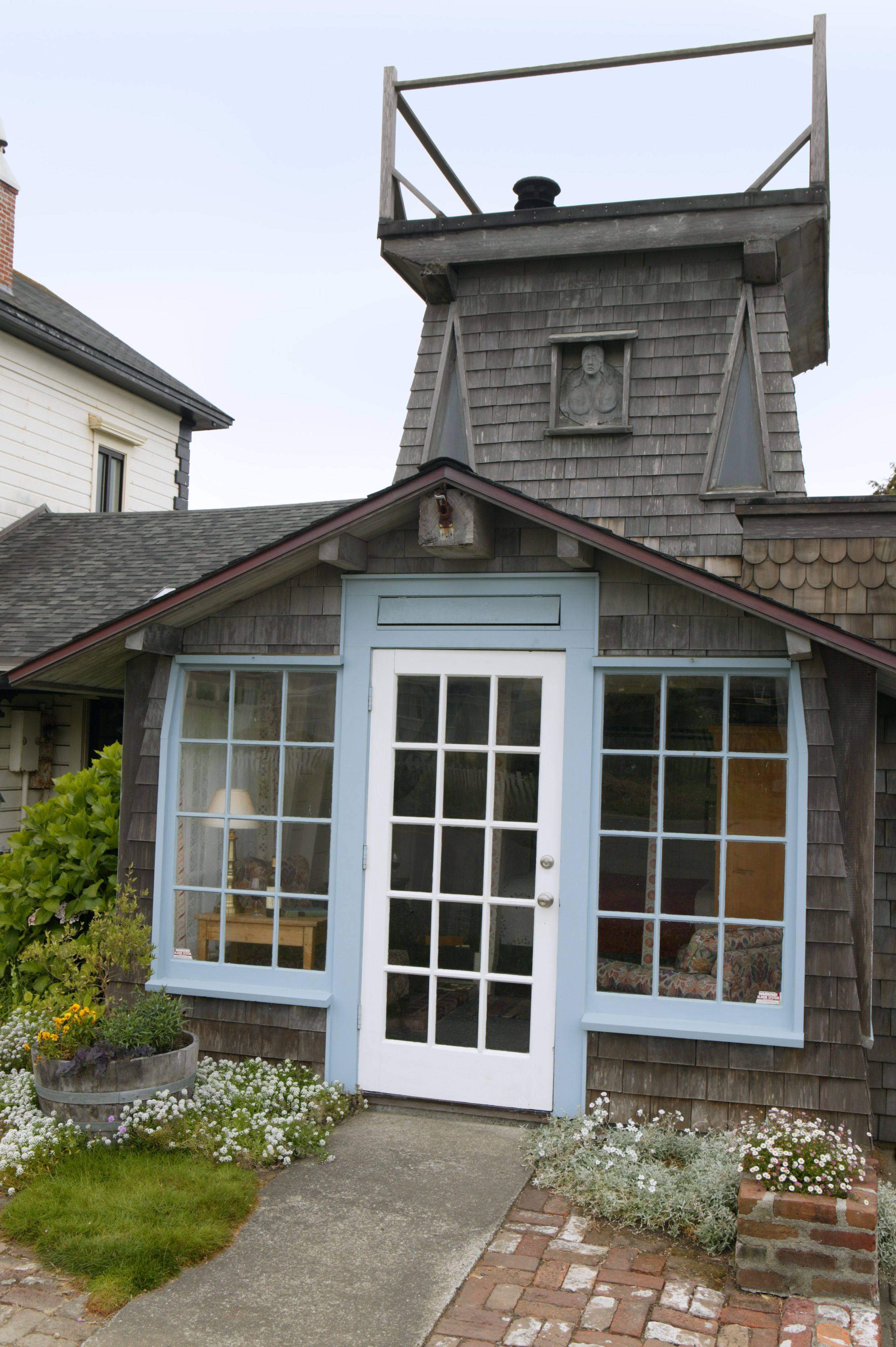 A small, light blue wooden cottage with a white door and windows, surrounded by a brick pathway and flower beds.