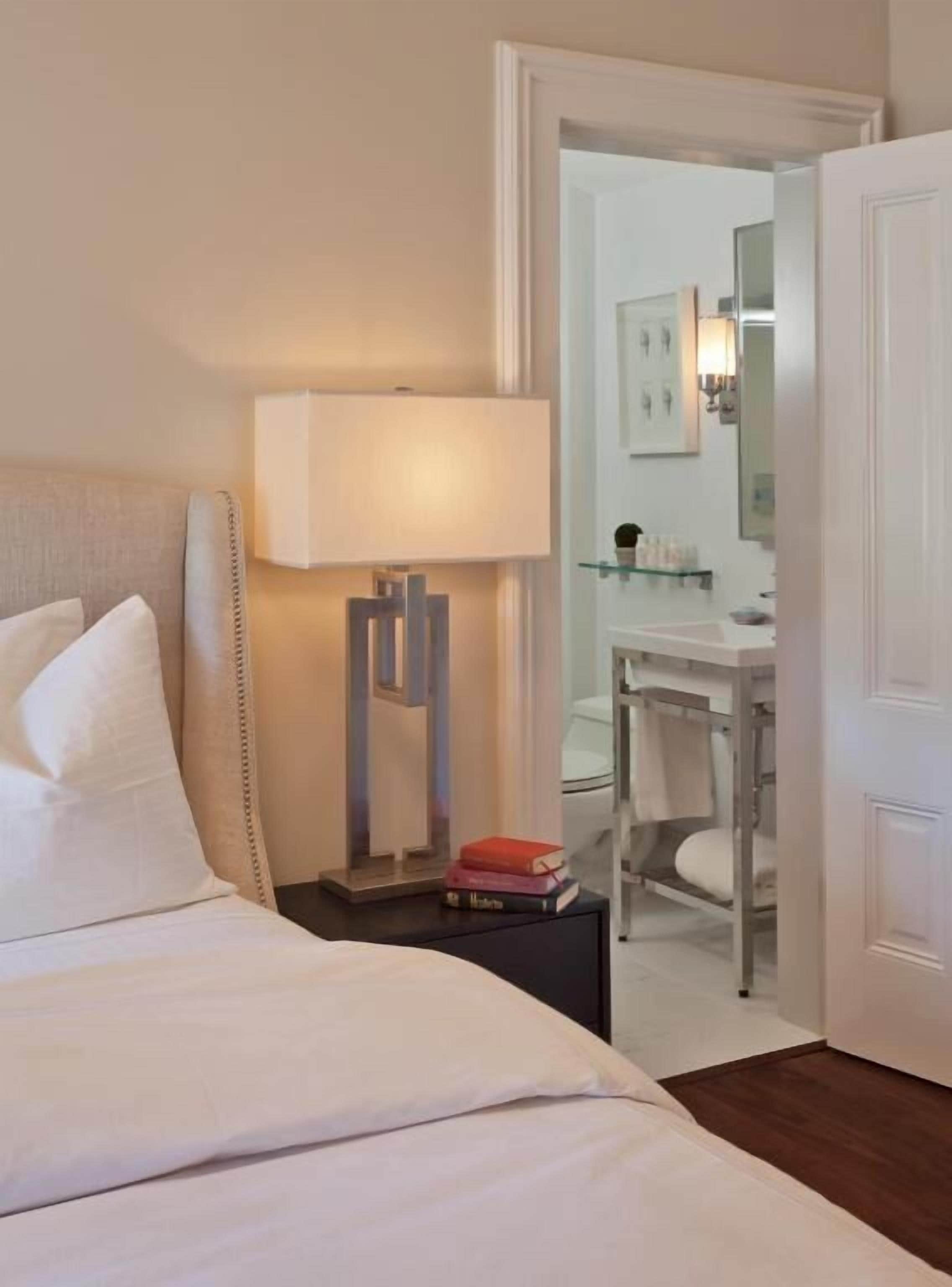 Habitación de hotel con una cama, una mesita de noche con lámpara, un escritorio pequeño y un baño visible a través de una puerta abierta.