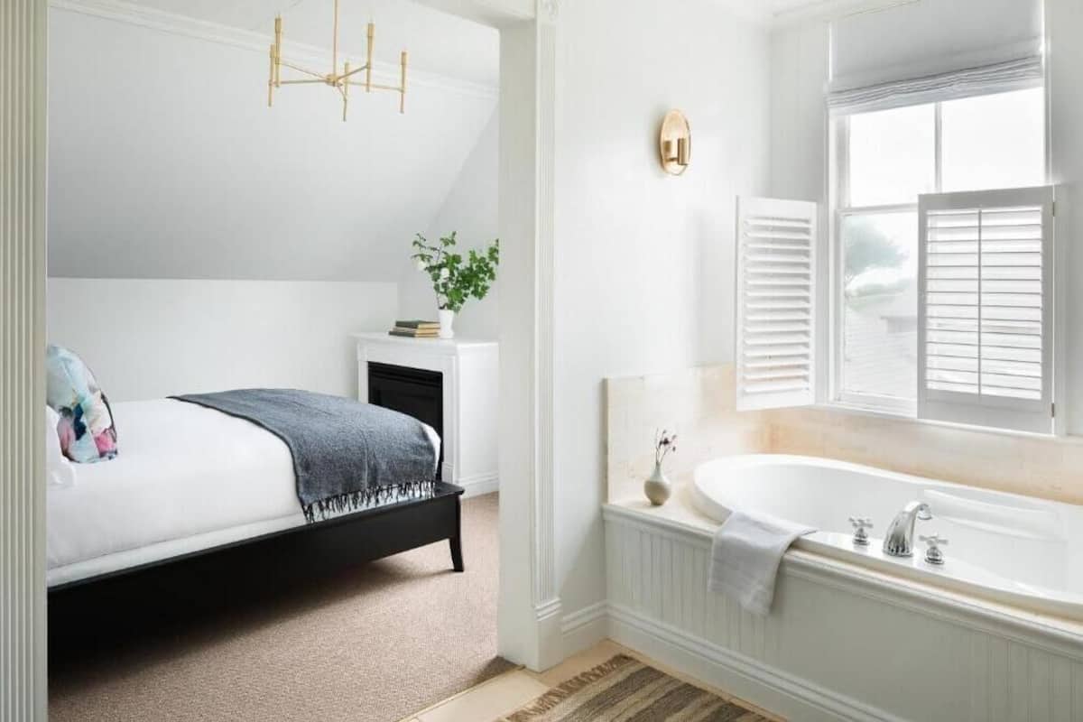 Un dormitorio con cama, una mesita de noche con una planta, una ventana con contraventanas y una bañera blanca.