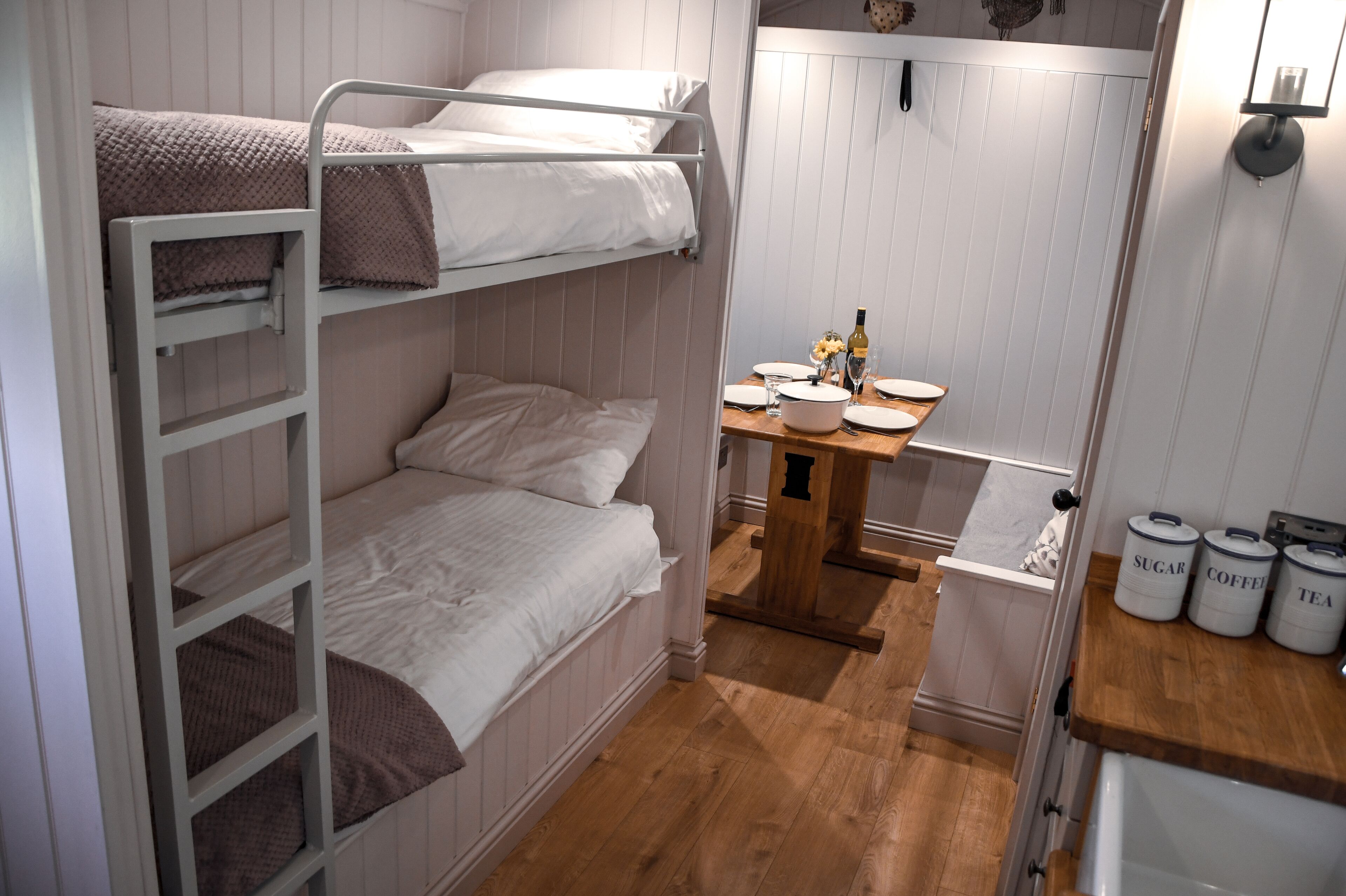 A small, compact room with bunk beds, a wooden table with a vase, and storage containers labeled 'SUGAR,' 'COFFEE,' and 'TEA.'