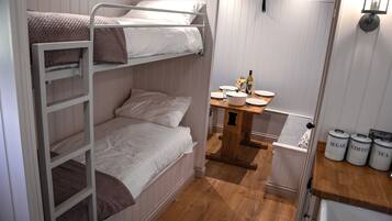 A small, compact room with bunk beds, a wooden table with a vase, and storage containers labeled 'SUGAR,' 'COFFEE,' and 'TEA.'