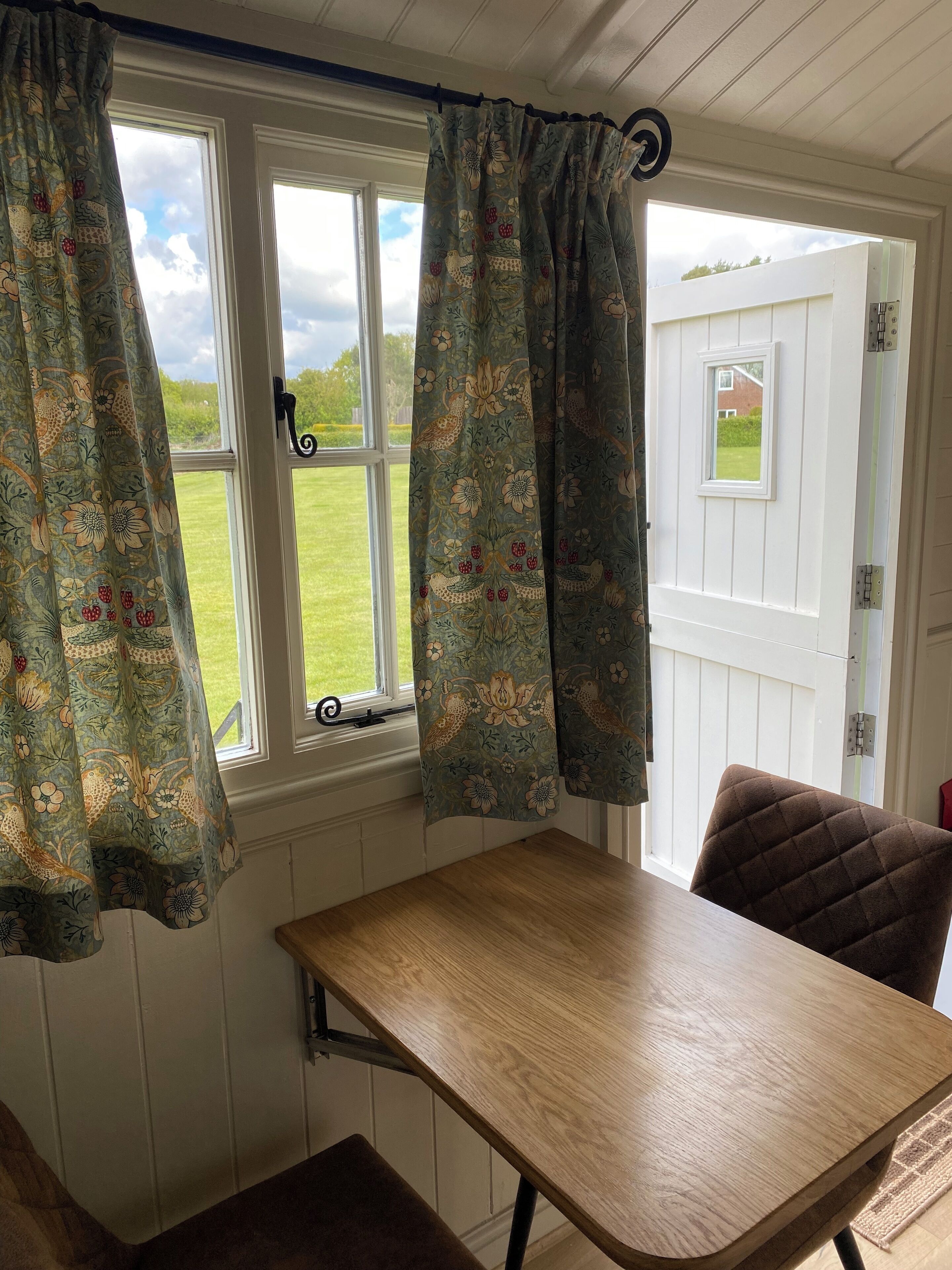 Una habitación con una mesa de madera y una silla, una ventana con cortinas florales y vistas a un césped verde.