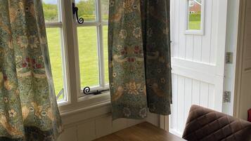 Una habitación con una mesa de madera y una silla, una ventana con cortinas florales y vistas a un césped verde.