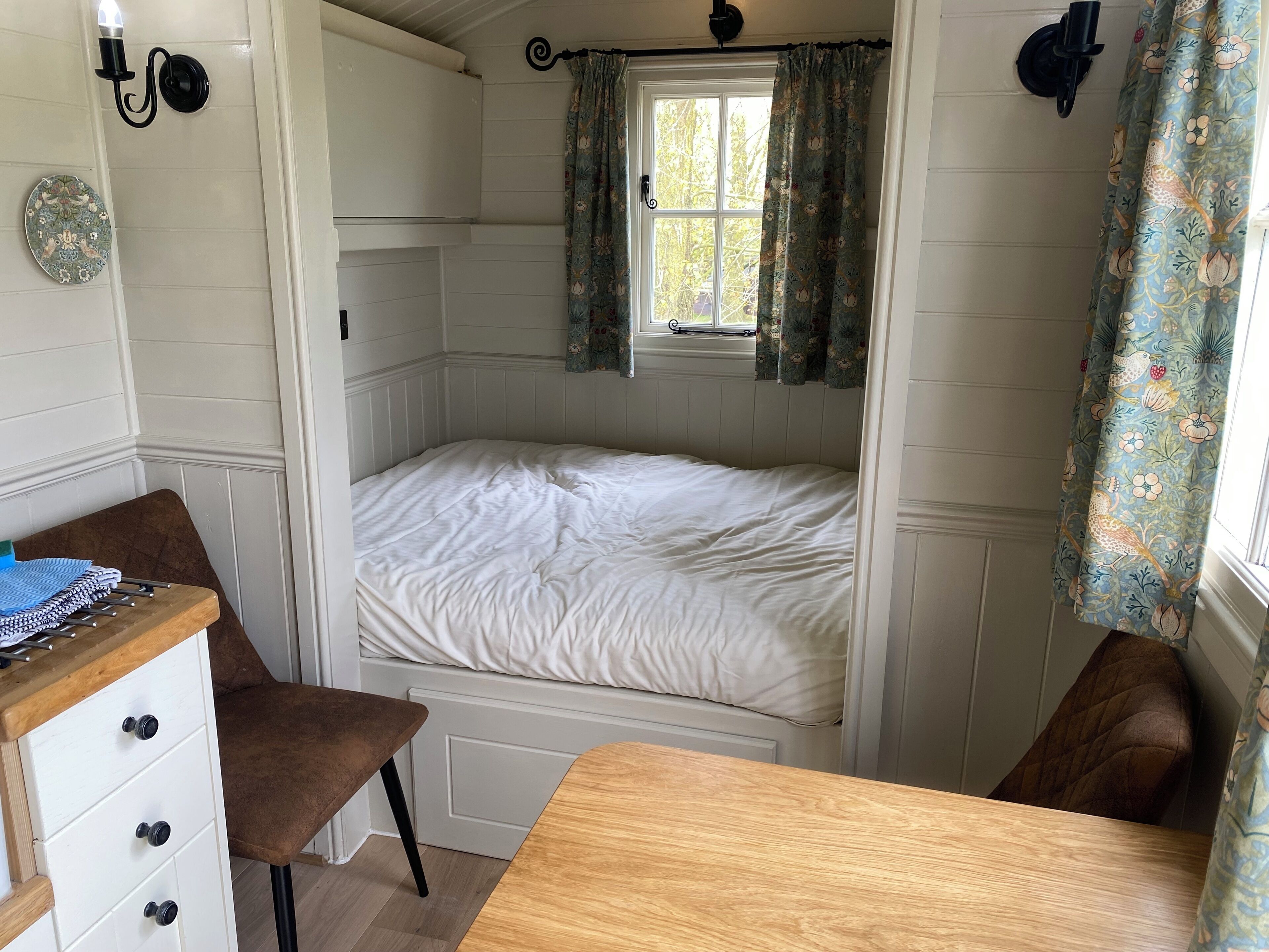 A small, cozy bedroom with a single bed, a wooden desk, a chair, and a window with floral curtains.